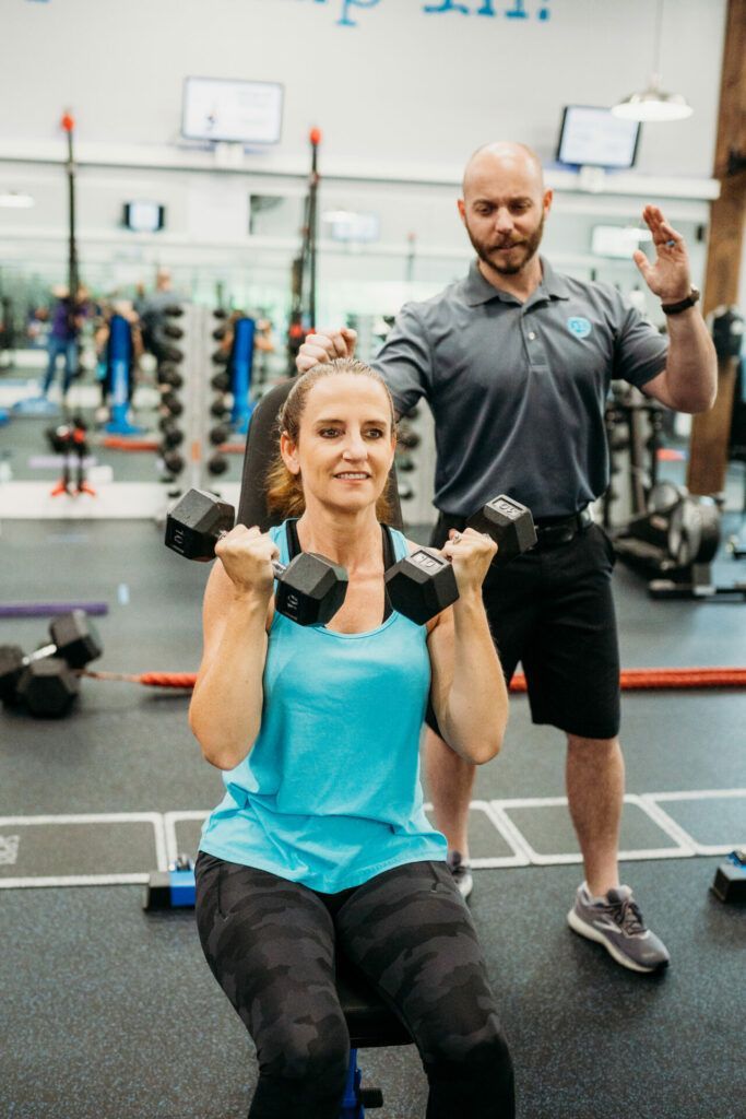Woman doing dumbbell curls, instructed by a trainer in a gym. She's wearing blue, sitting. He wears gray.