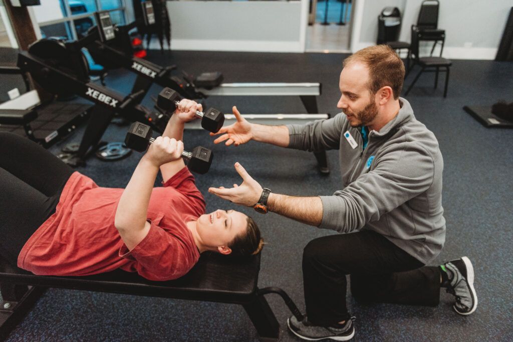 Woman on bench lifting dumbbells, assisted by trainer in gym.