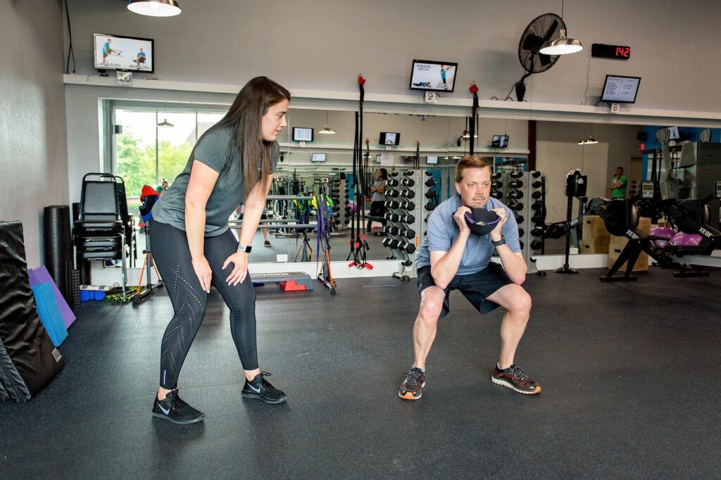 A fitness instructor watches a man squat while holding a kettlebell in a gym.