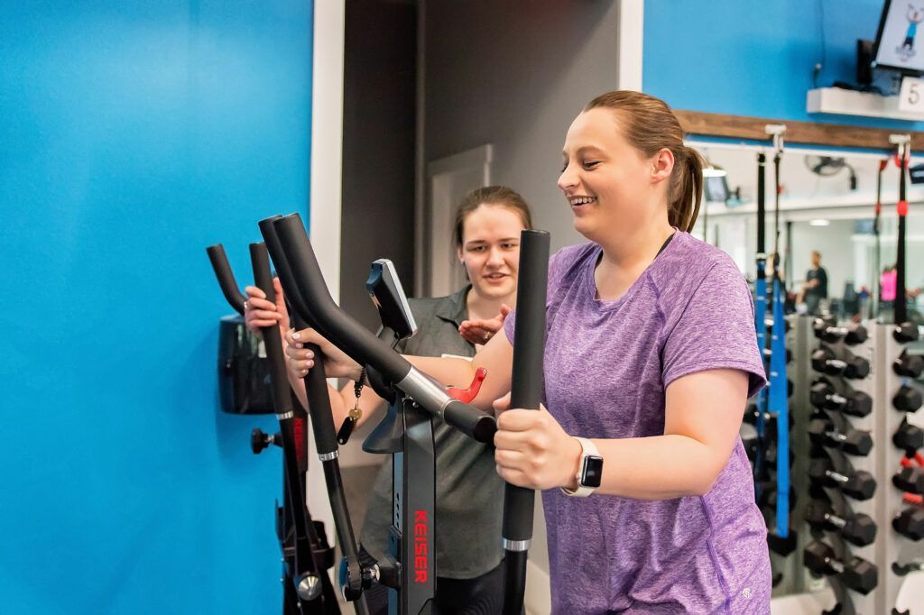 Two women using elliptical machines in a gym. One smiles, wearing a purple shirt. Blue wall in the background.
