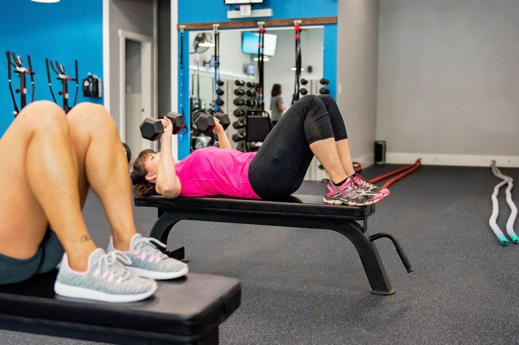 Woman doing dumbbell chest press on a bench in a gym. Another person sits on a bench in foreground.
