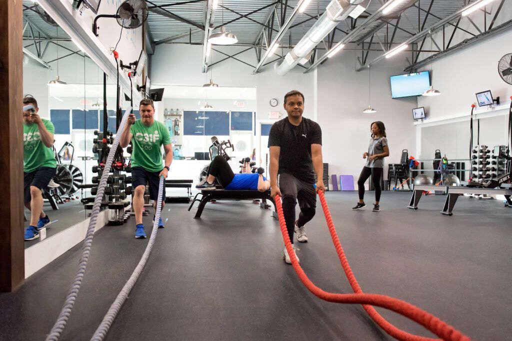 People exercising with ropes in a gym; gray floor, mirrors, weights, and blue sky visible outside.