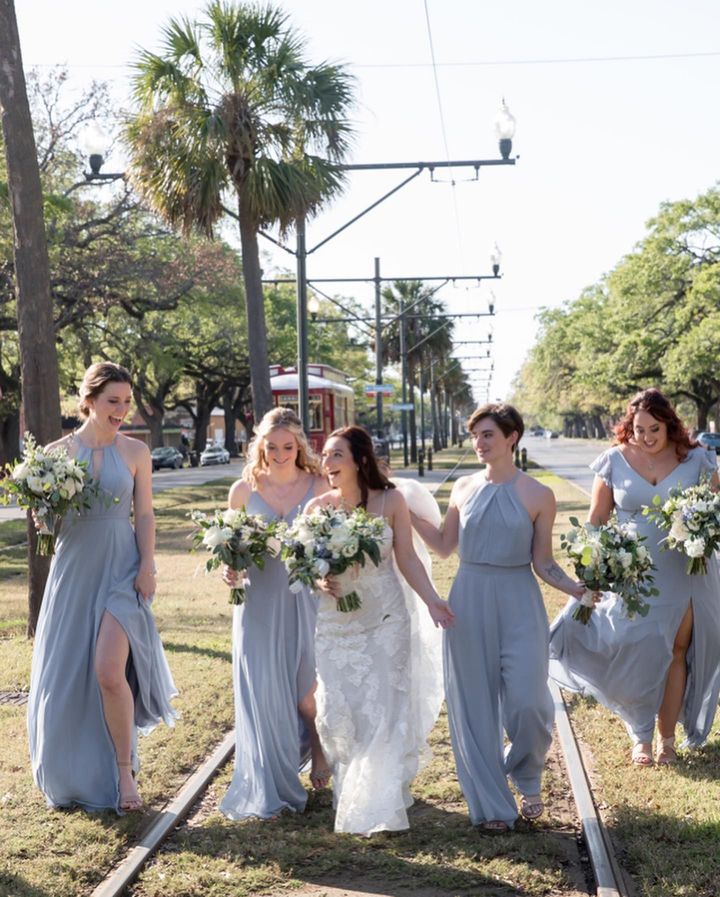 Bride and bridesmaids walking along train tracks, holding bouquets, wearing blue dresses, laughing.