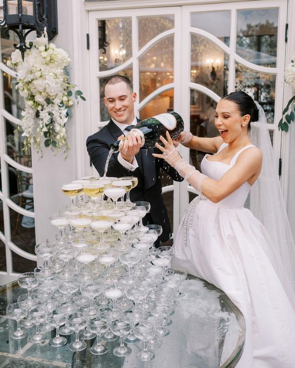 Bride and groom pouring champagne into a tower of glasses, celebrating at an outdoor wedding.