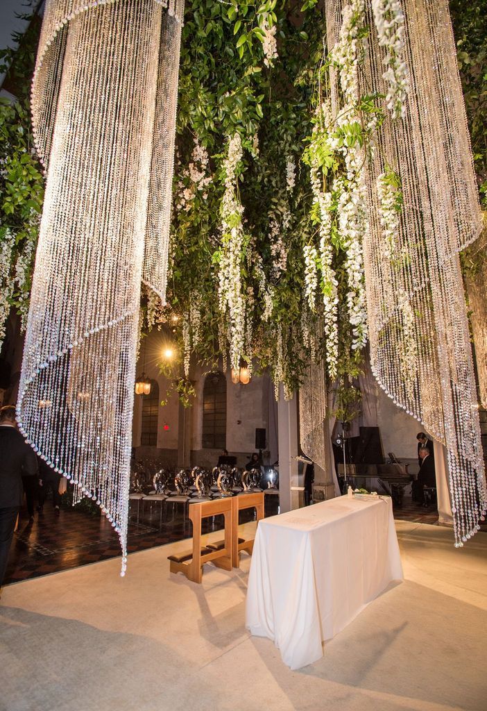 A table with a white table cloth is sitting under a tree with flowers hanging from it.