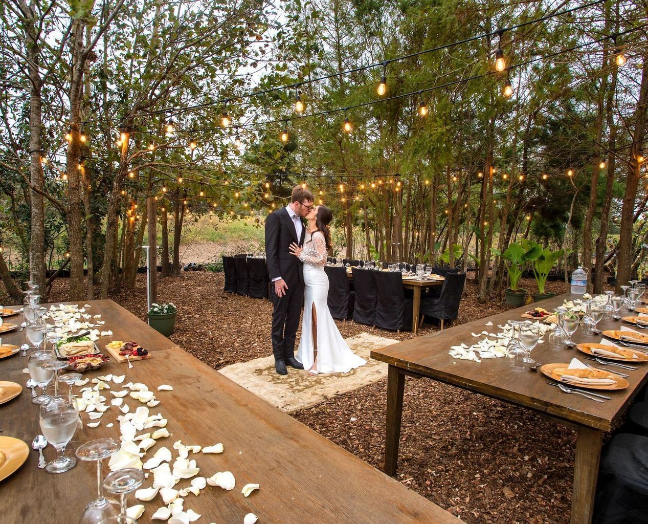 A bride and groom are kissing in front of a long wooden table.