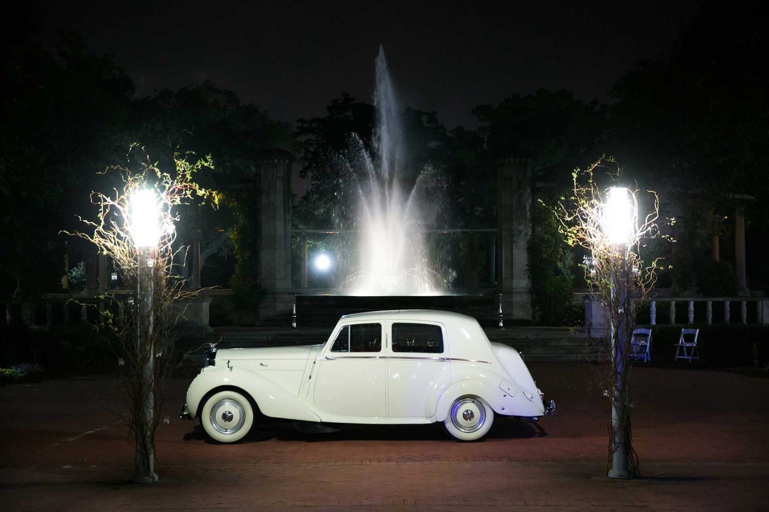 A white car is parked in front of a fountain at night.