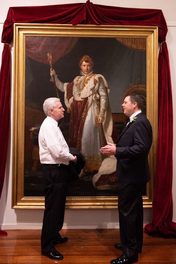Two men are standing in front of a large painting of a king.