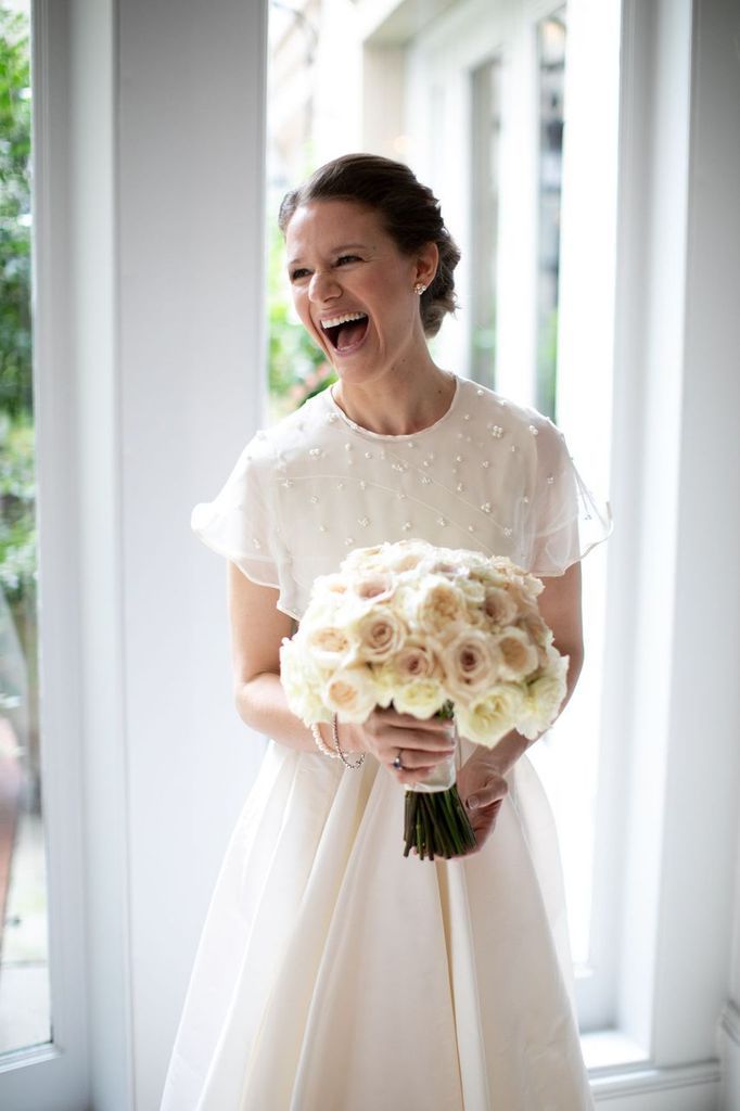 A bride in a white dress is holding a bouquet of white flowers and laughing.
