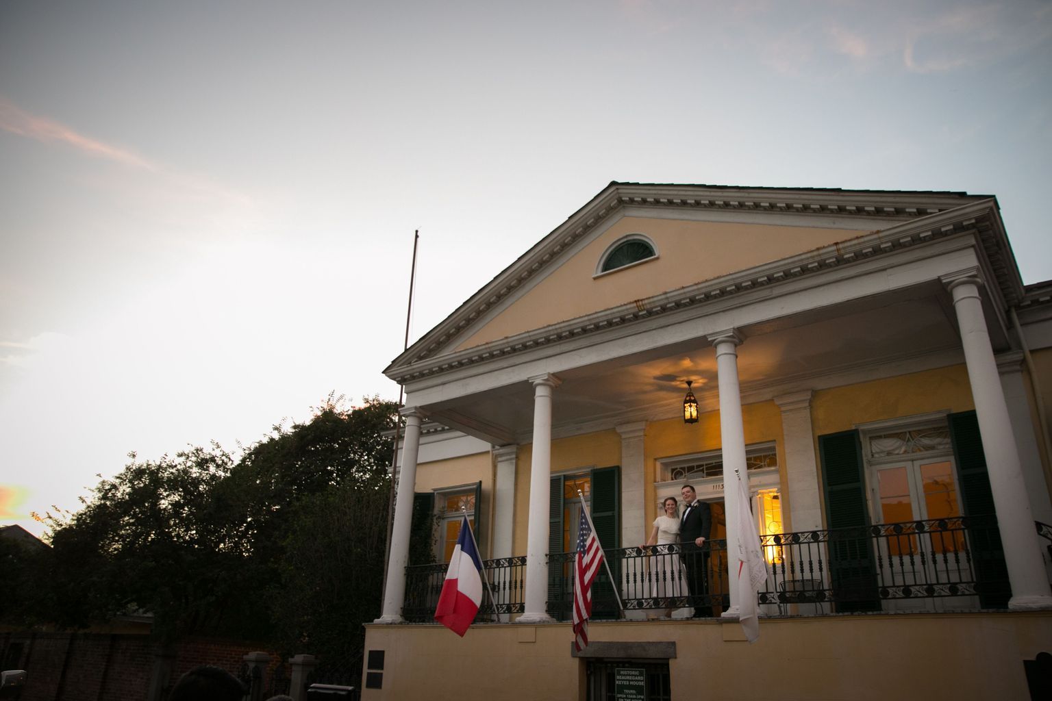A man and woman are standing on a porch of a building with a french and american flag.