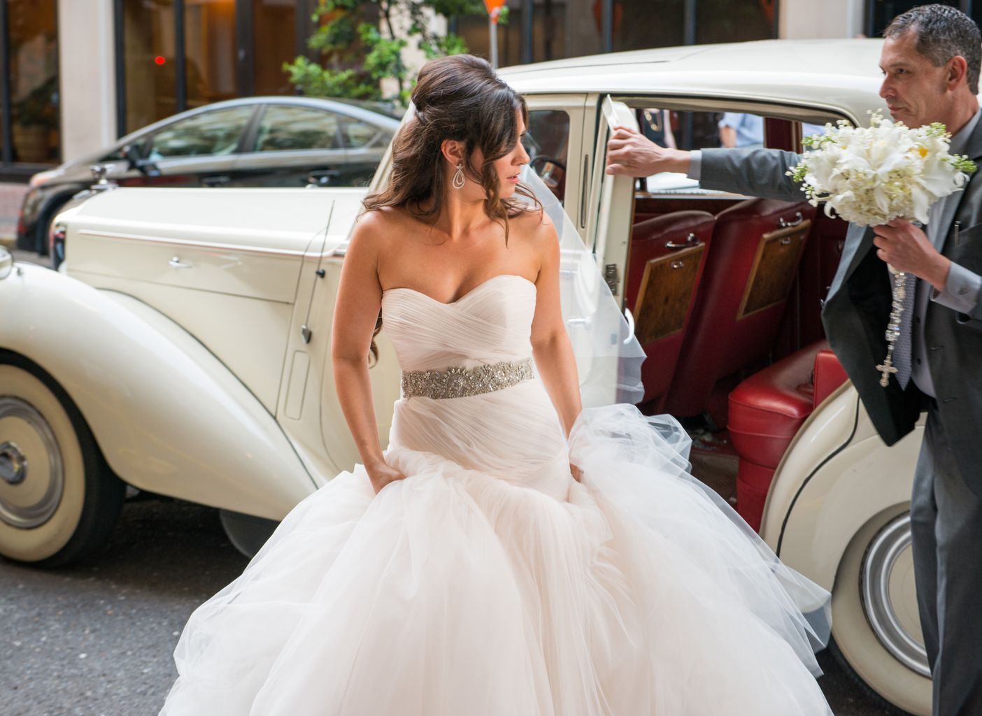 A bride in a wedding dress is getting out of a white car.
