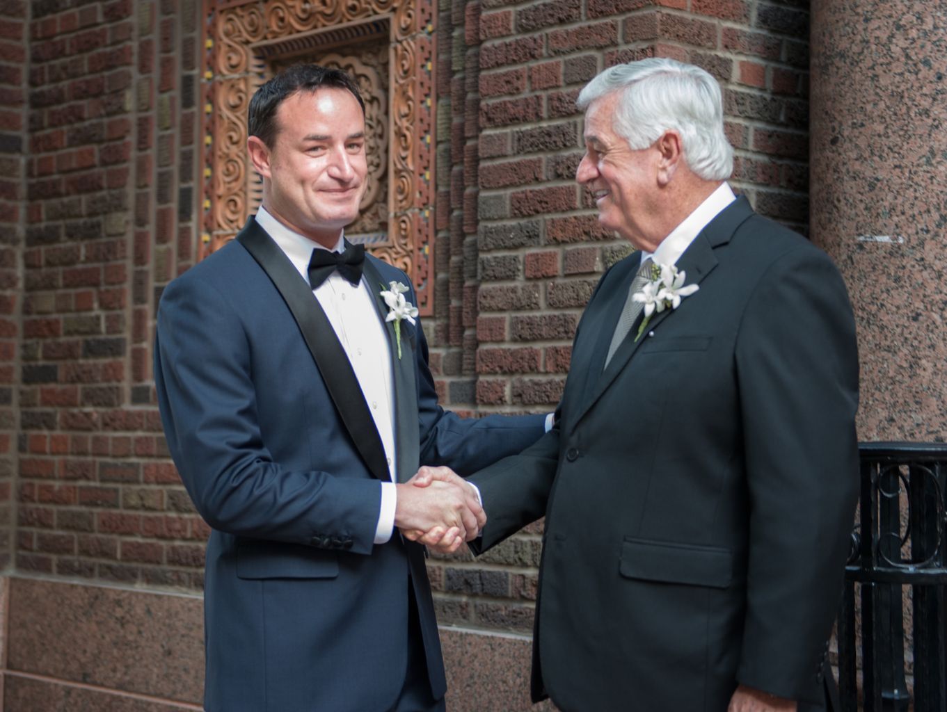 Two men in suits are shaking hands in front of a brick building.