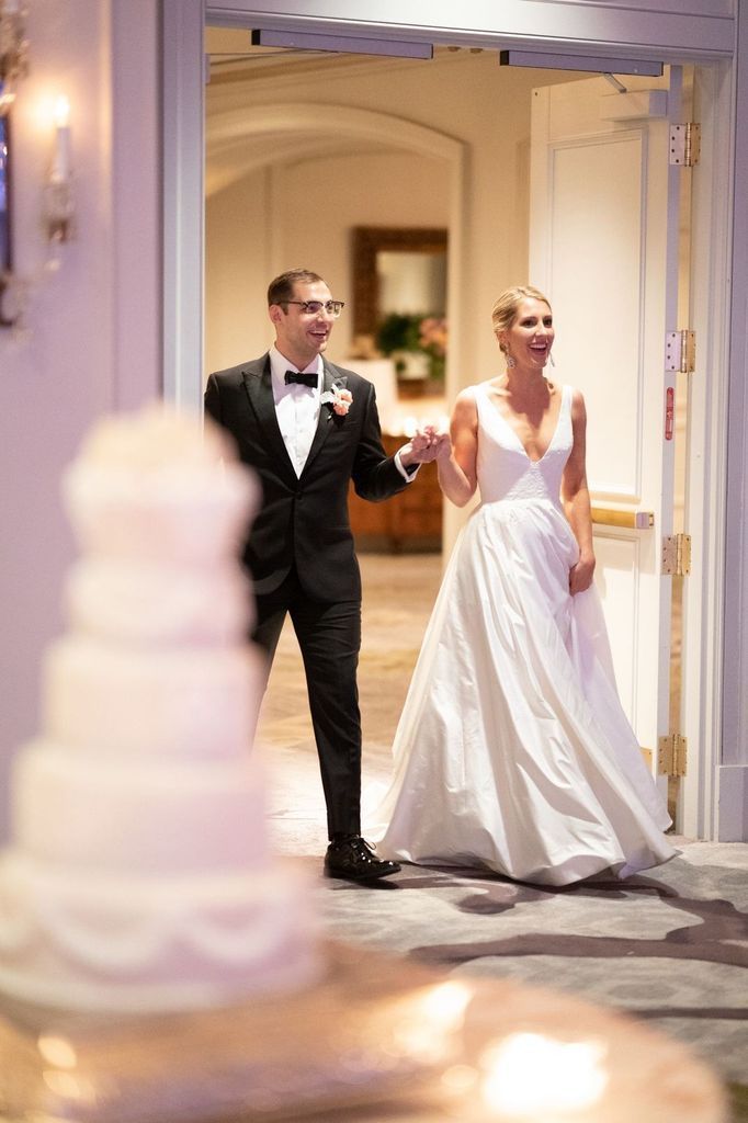 A bride and groom are walking into a room with a cake in the background.