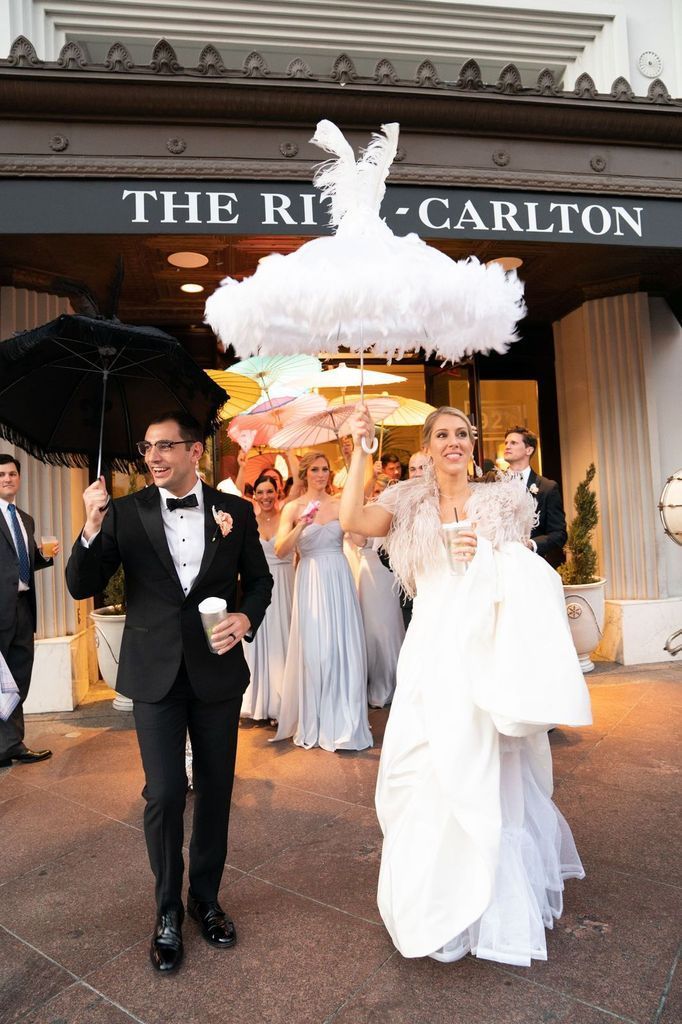 A bride and groom are walking with umbrellas in front of a building.
