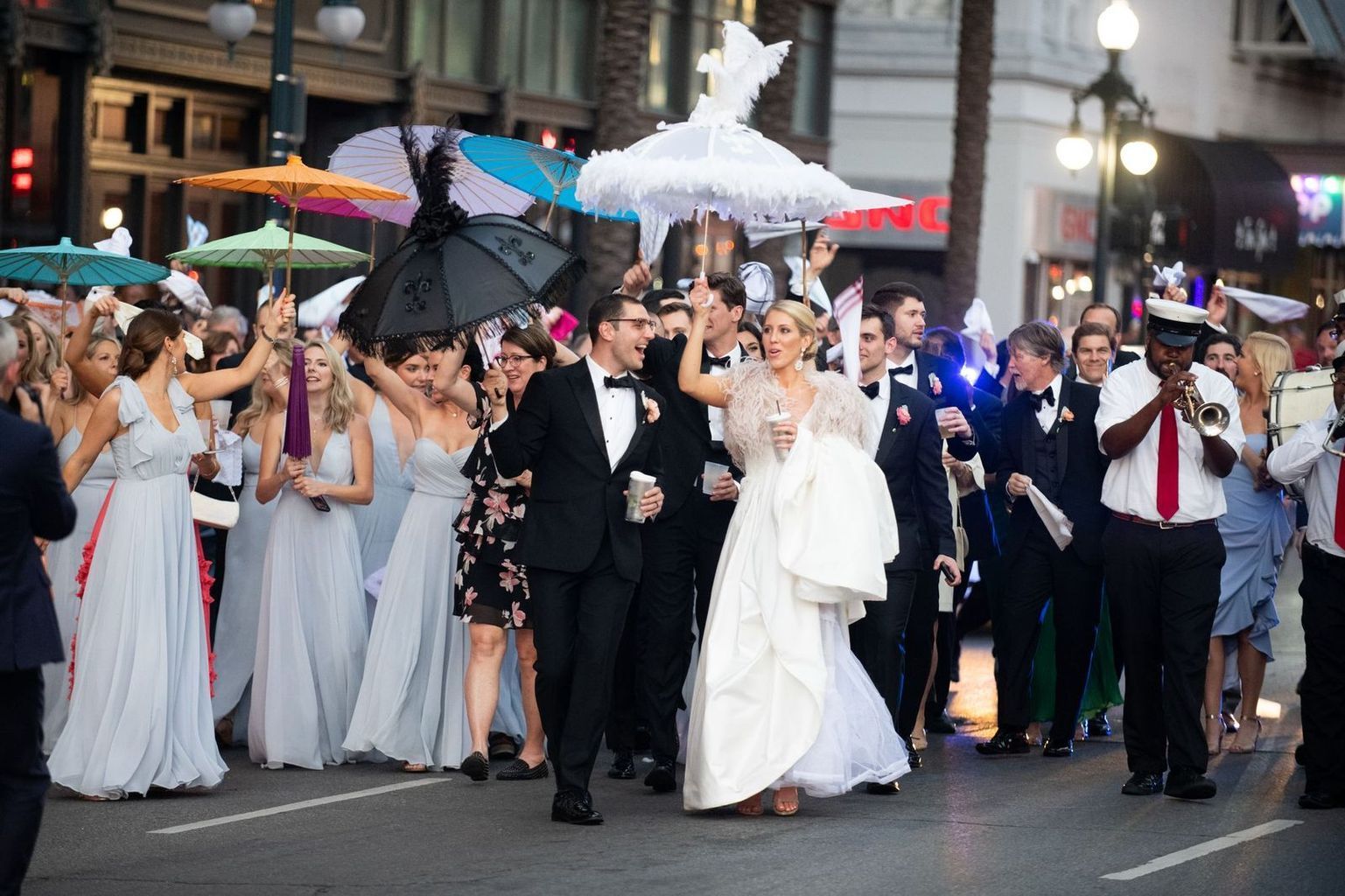 A bride and groom are walking down the street with their wedding party holding umbrellas.