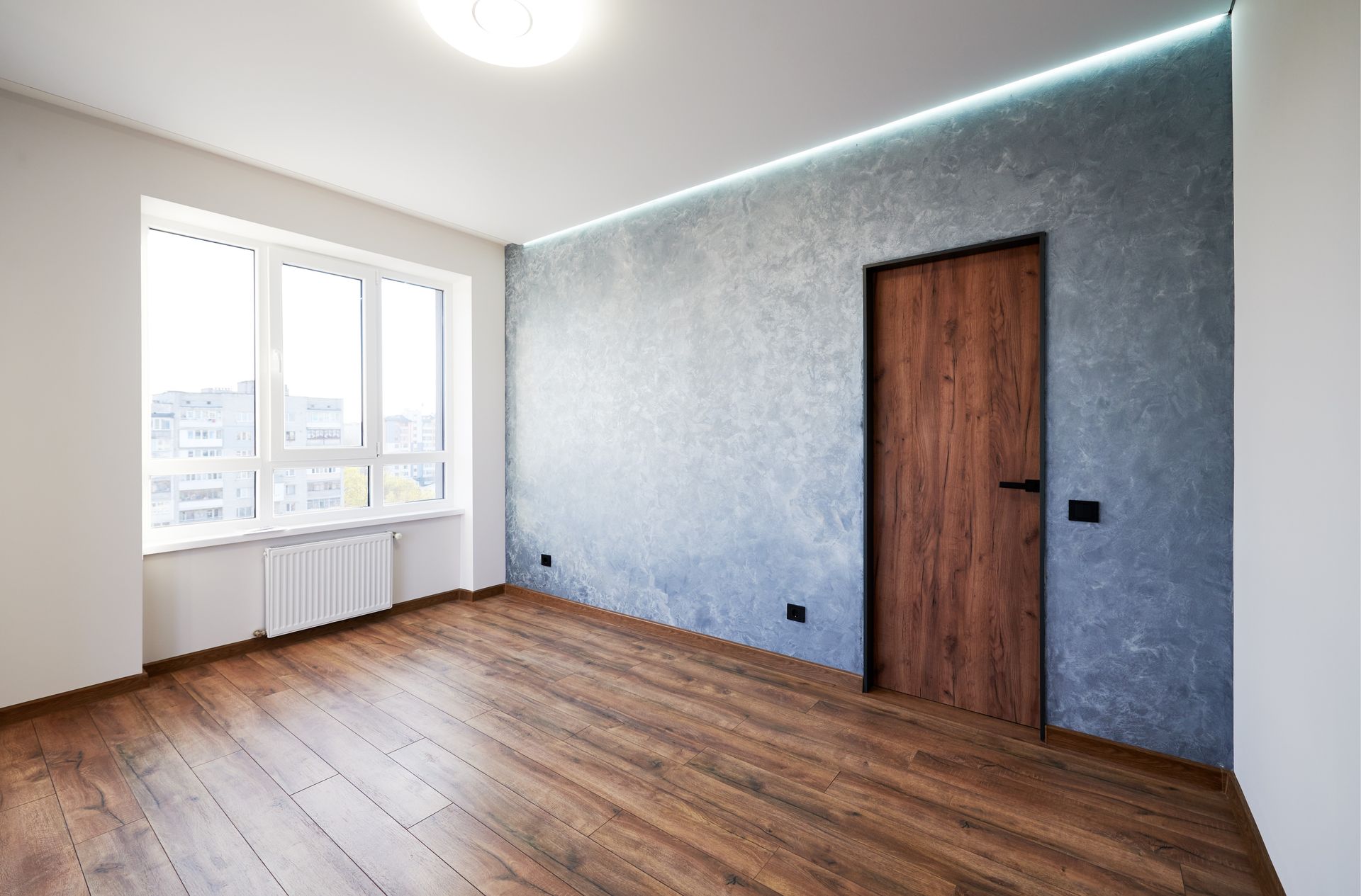 Empty room with wood flooring, a window, and a textured blue wall with a wooden door.