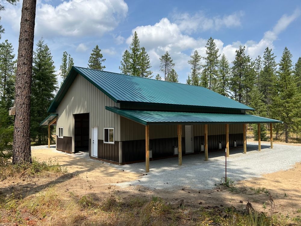 Tan and brown barn with a teal roof and porch, set in a clearing surrounded by pine trees.