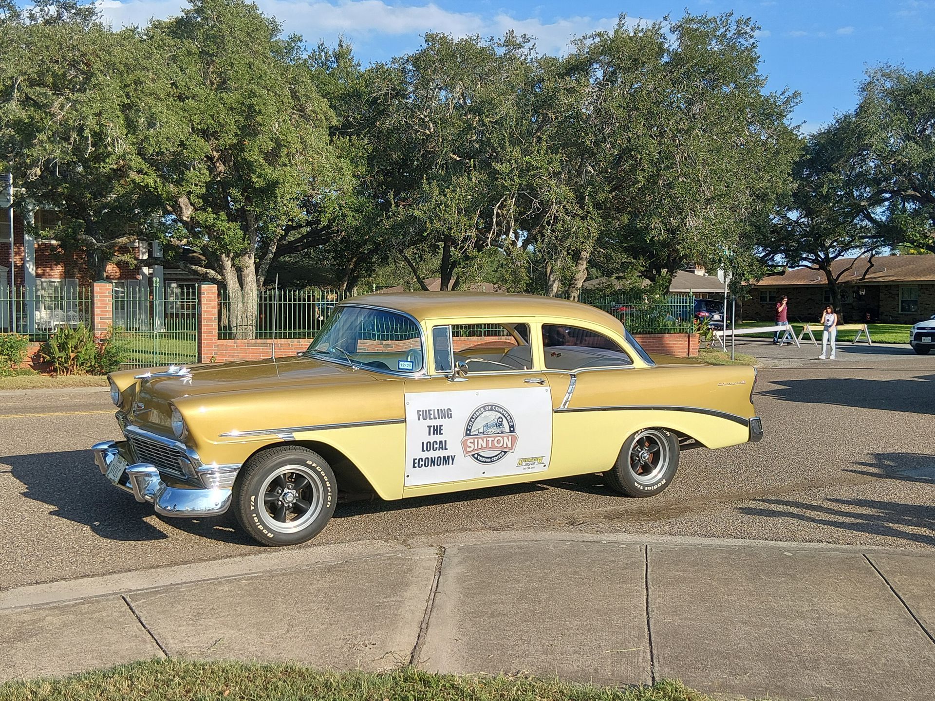 Gold classic car parked on a paved driveway with a sign on the door, trees in the background.