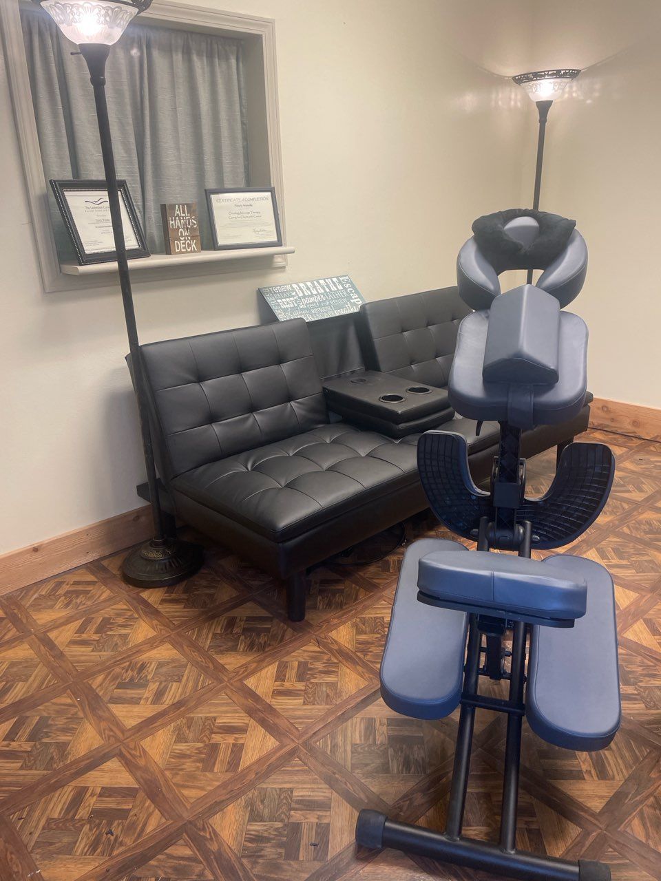A massage chair stands in a room with a black futon, two floor lamps, and diplomas on a window ledge.