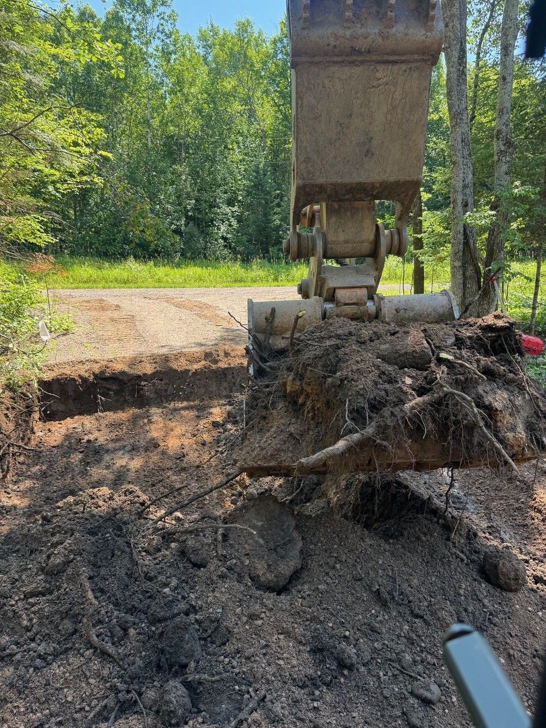 Excavator bucket scooping dark soil on a dirt road, trees in the background under a sunny sky.