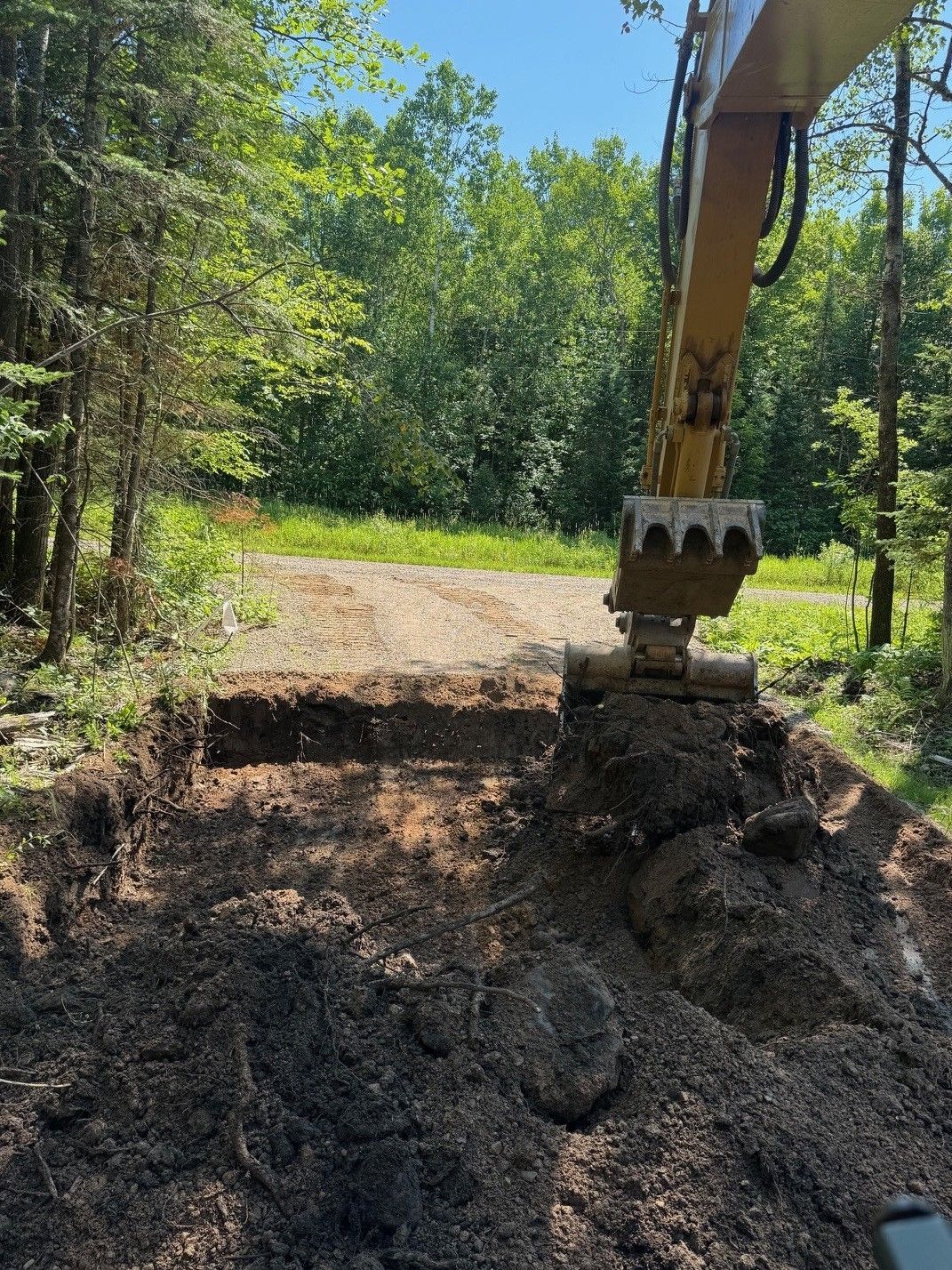 Excavator digging into the side of a gravel road, with trees visible in the background on a sunny day.
