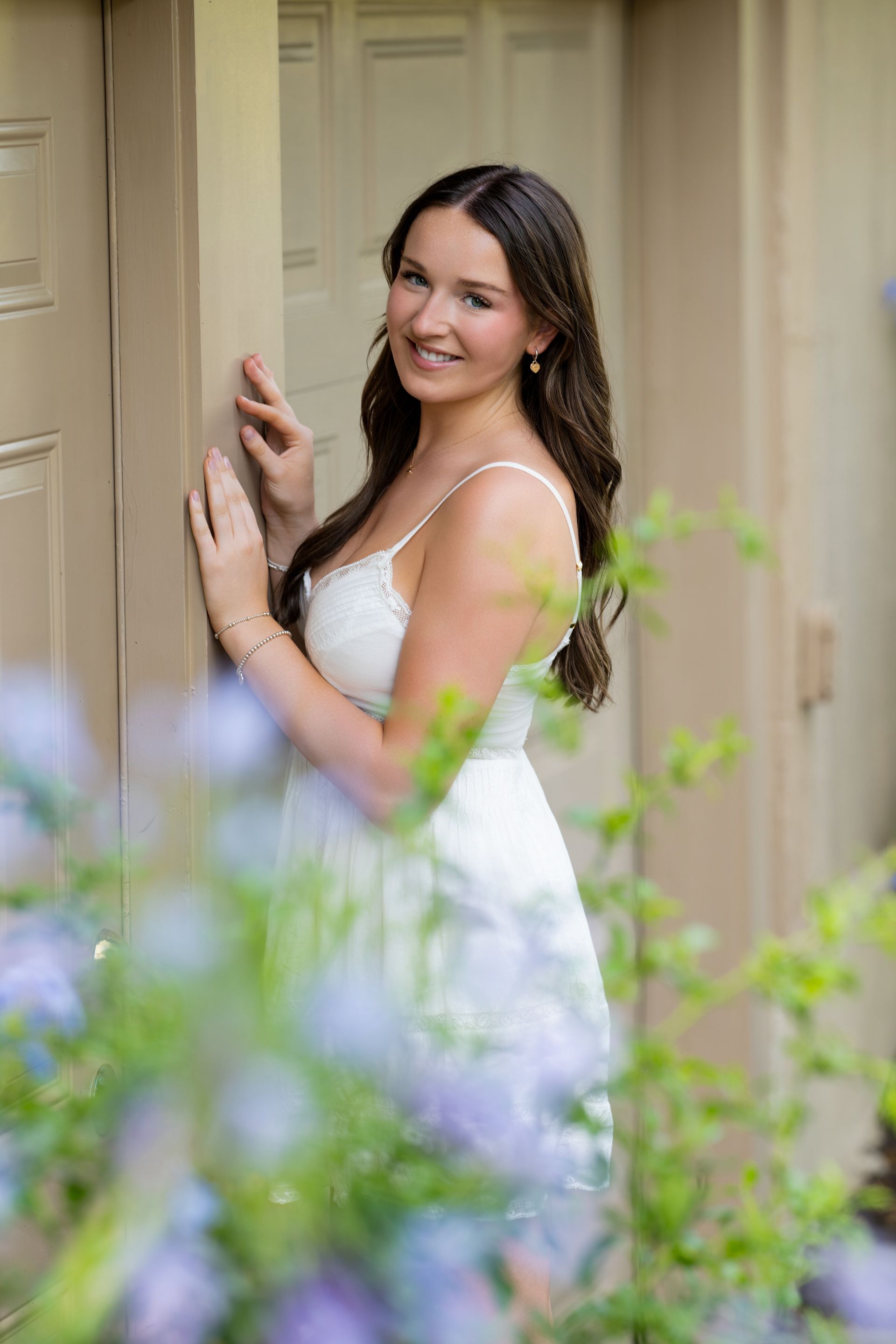 Woman in a white dress smiling by a doorway with soft greenery in the foreground