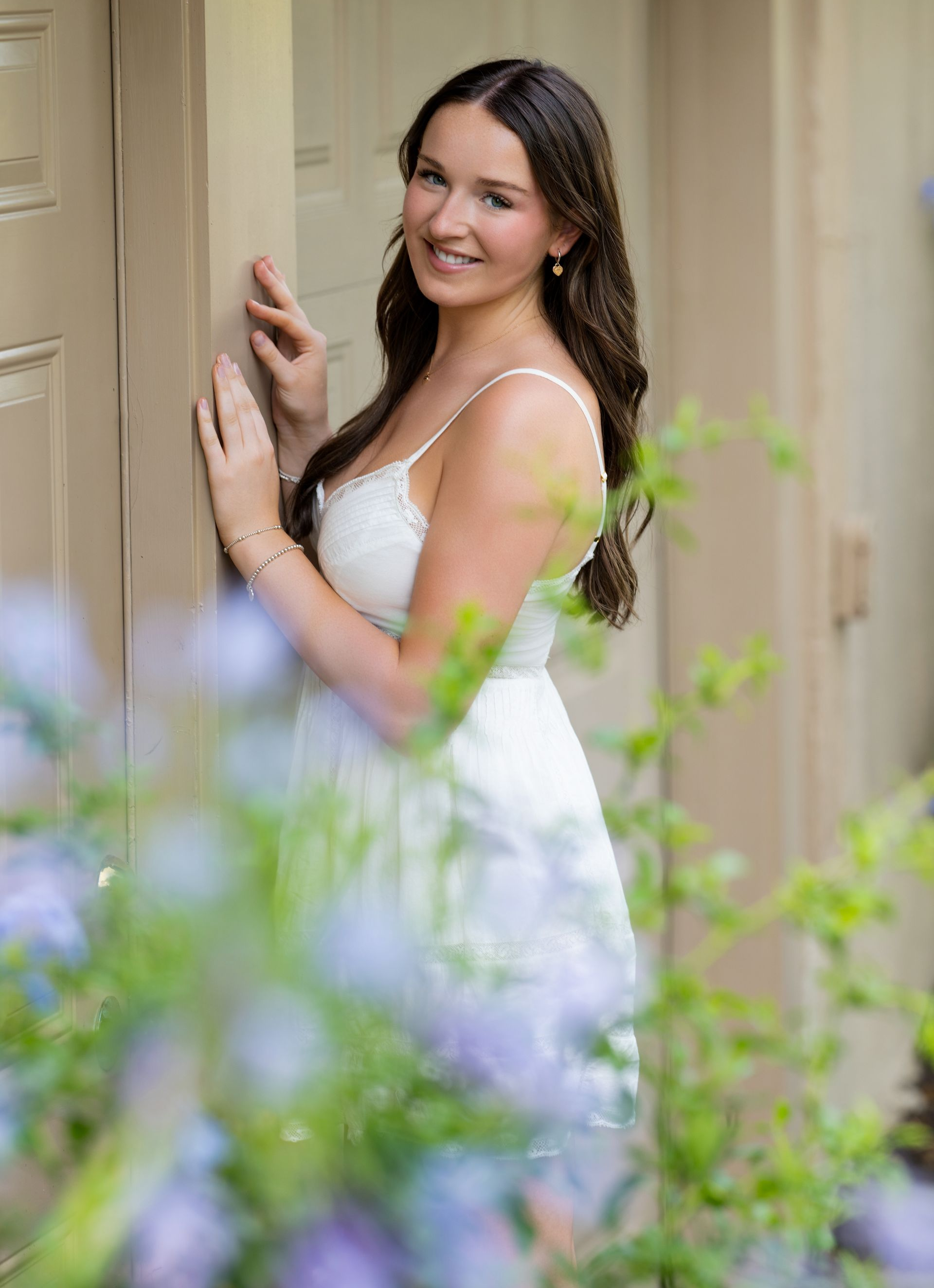Woman in a white dress smiling by a doorway with soft greenery in the foreground