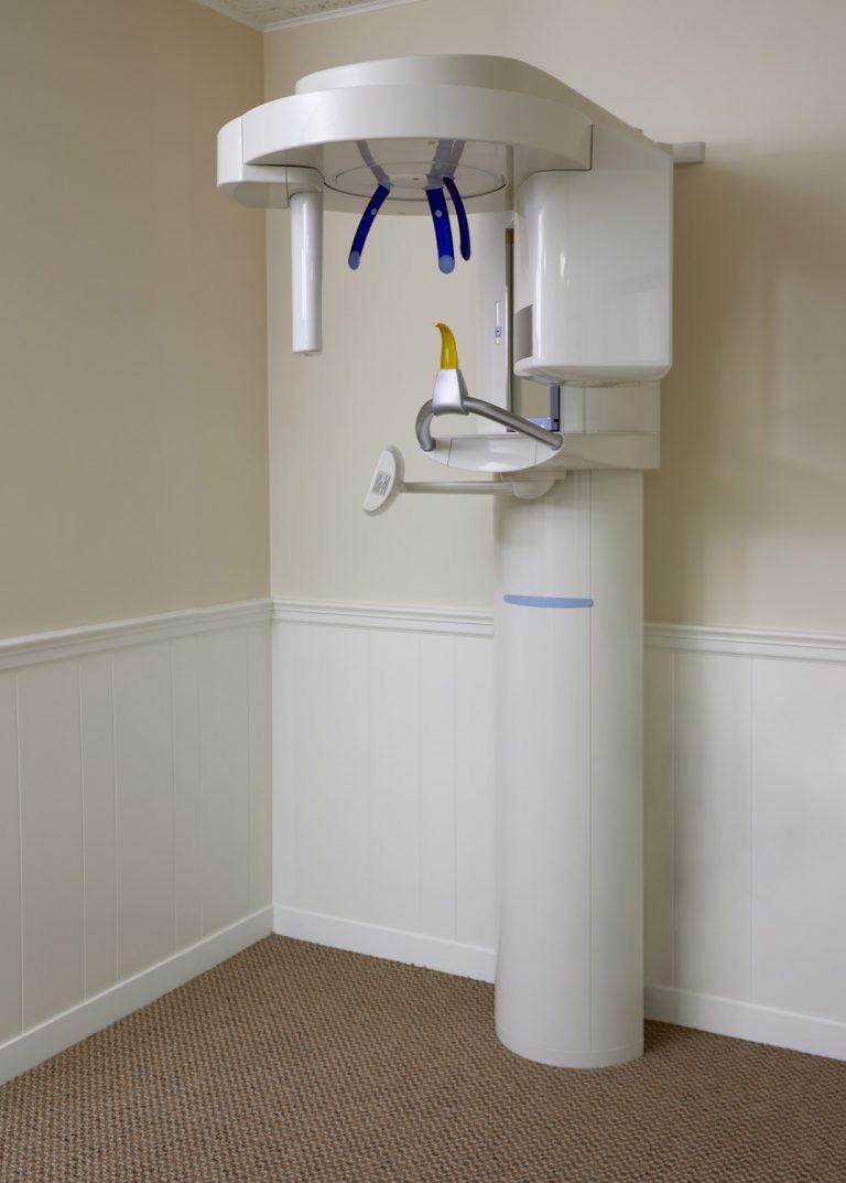 Dental X-ray machine in a treatment room, with arm and head positioned over a chair space.