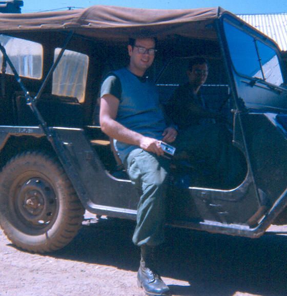 Man leaning against a black jeep with a tan canvas top, holding a drink outdoors in bright sunlight