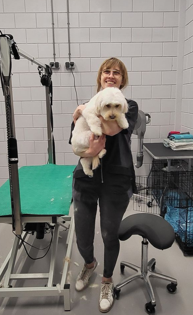 Een vrouw houdt een pluizige witte hond vast in een trimsalon. Ze lachen allebei.