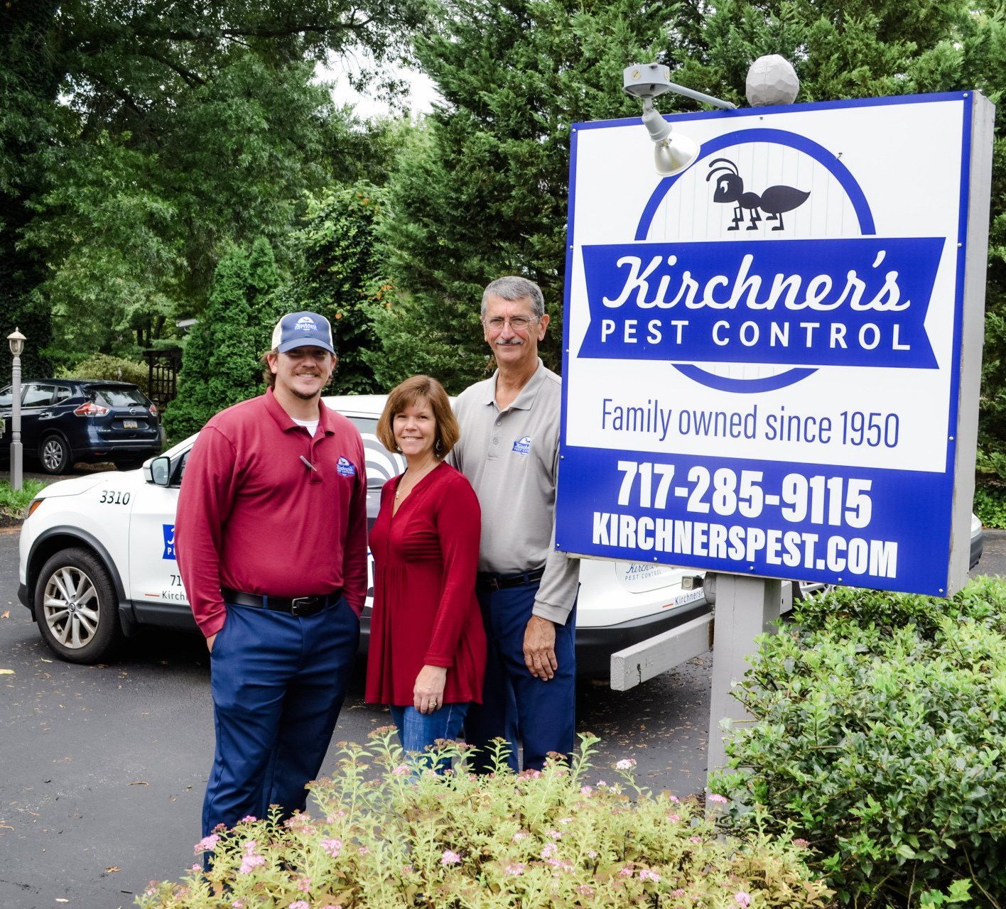 Three people stand by a Kirchner's Pest Control sign. A man in red leans against a white SUV. Two people stand in front of the sign.