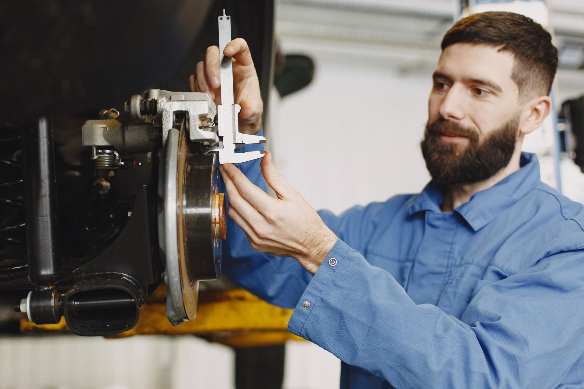 Mechanic in blue overalls measures a car's brake disc with calipers in a garage.