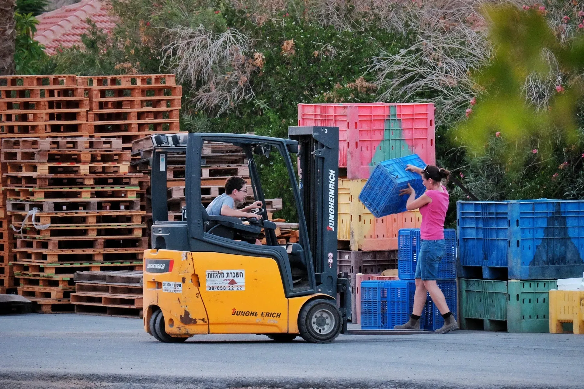 A woman operating a yellow forklift while another stacks blue crates outside near pallets.