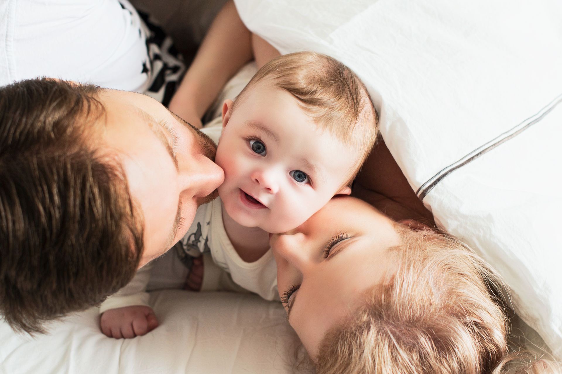 Three people lying together on a white bed, with a baby in the center looking up at the camera.