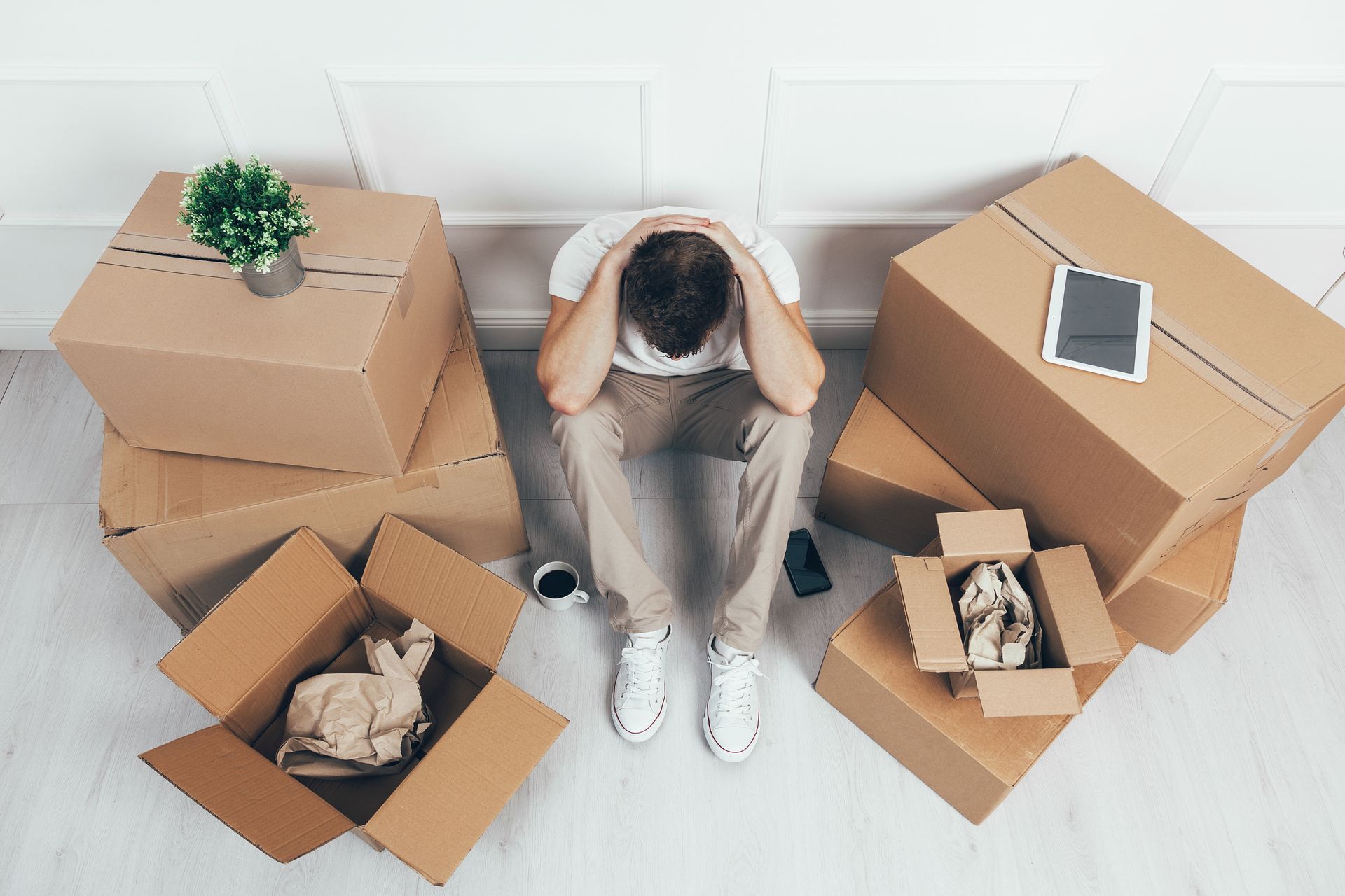 Person sitting on the floor among open moving boxes, head in hands, with a plant and tablet nearby.