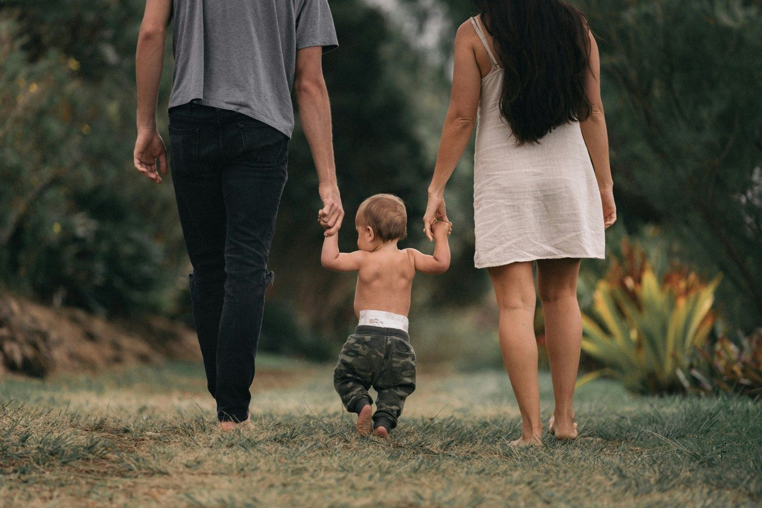 Three people walking hand in hand on a grassy path, with a toddler between two adults.