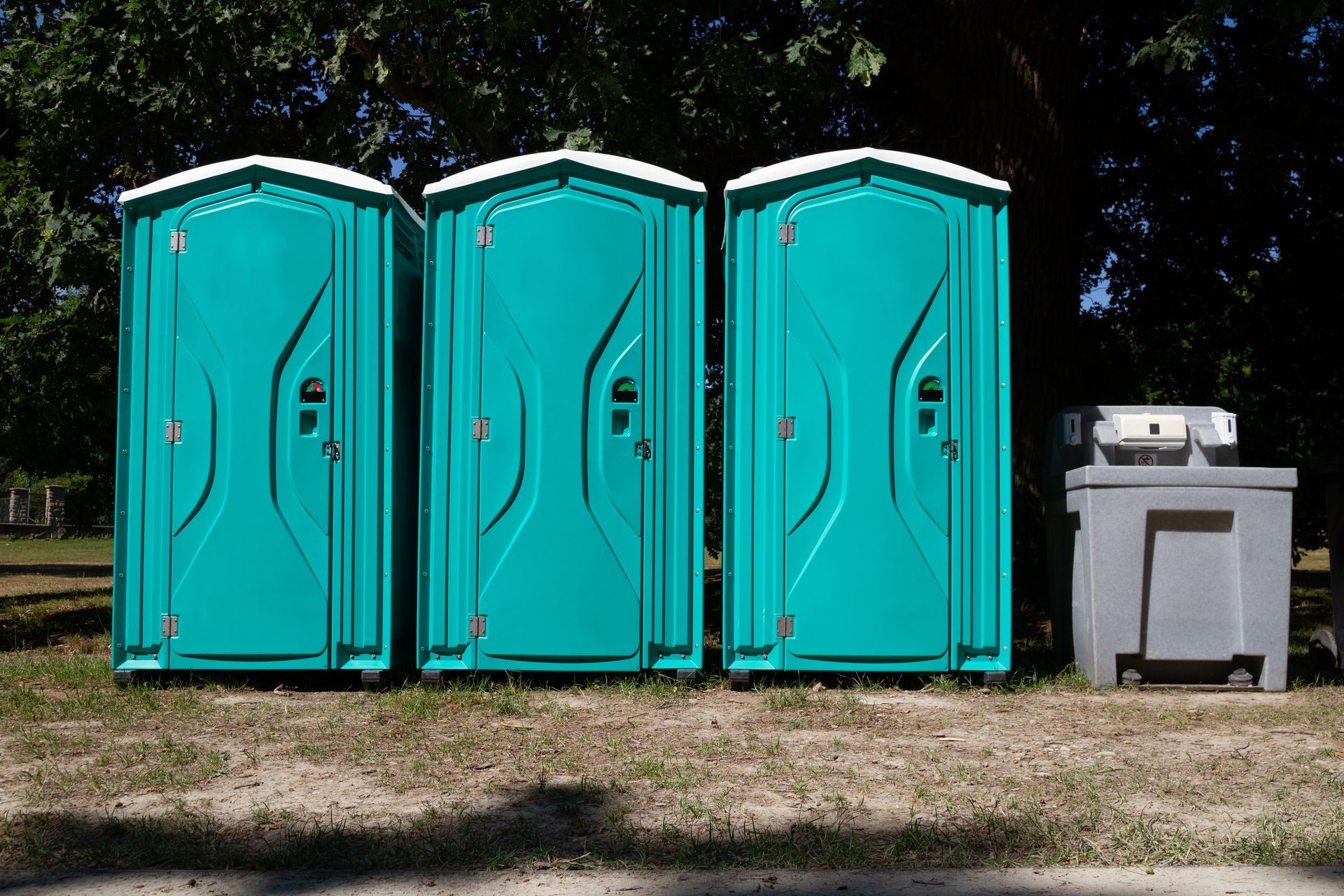Three teal portable toilets and a grey handwashing station in a sunny outdoor setting.