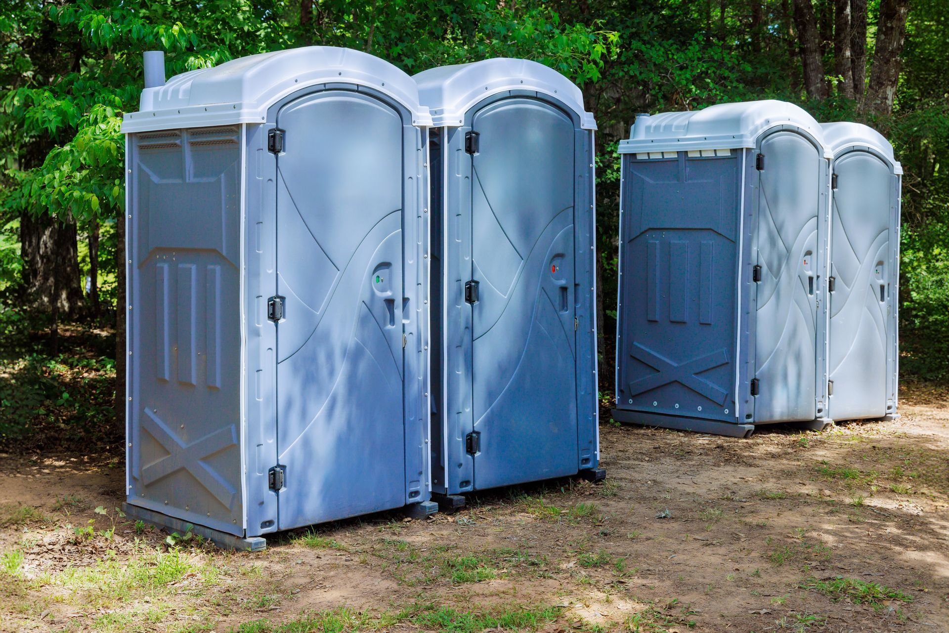 Four gray portable toilets lined up outdoors, set against a backdrop of trees.