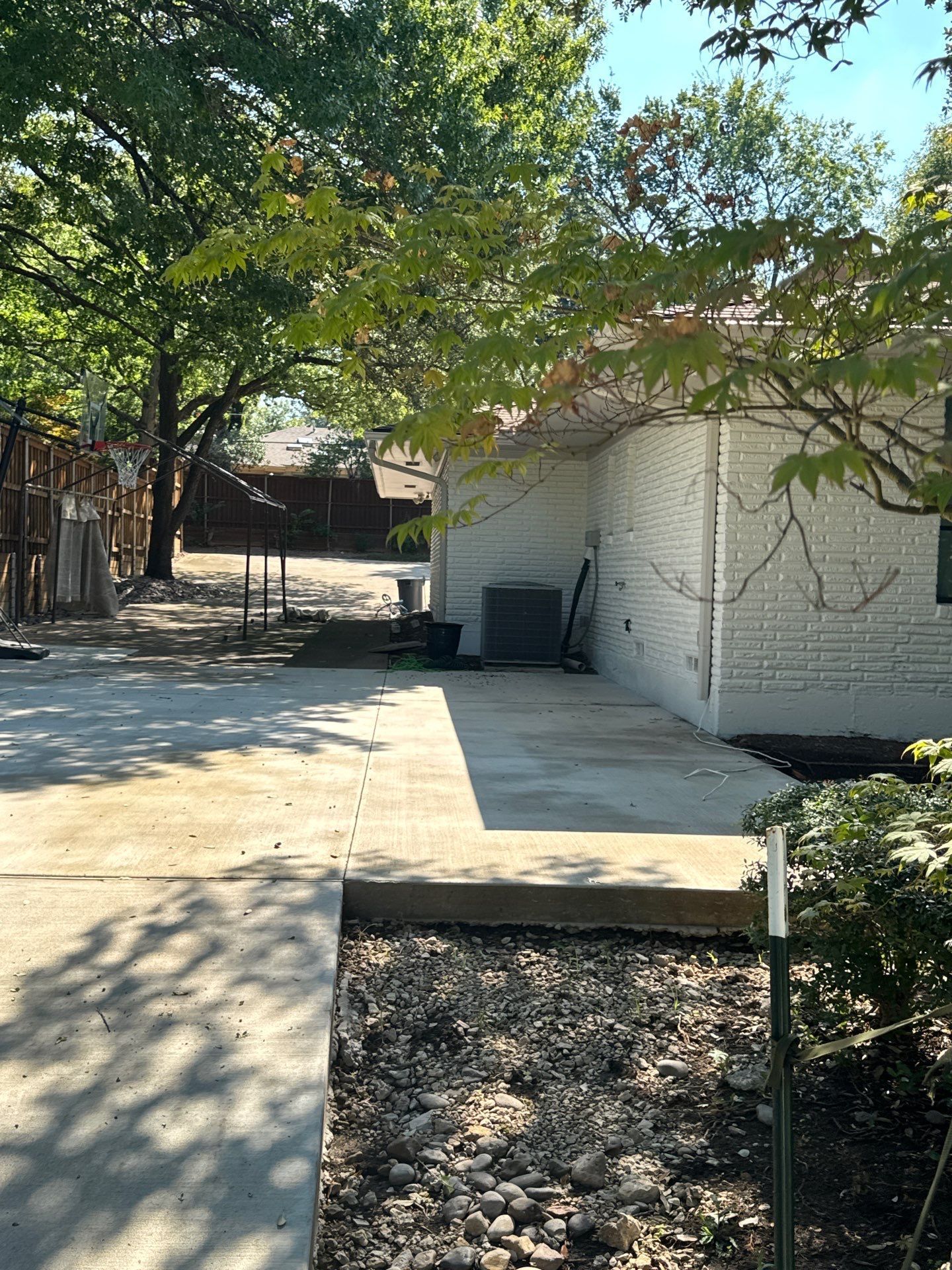 A person digs a trench in a backyard near a small excavator, a tree, and a house.