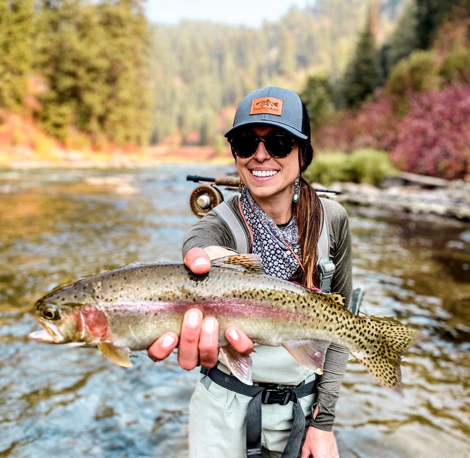 Woman holding fish and flyfishing rod in waders on the river with forest in the background