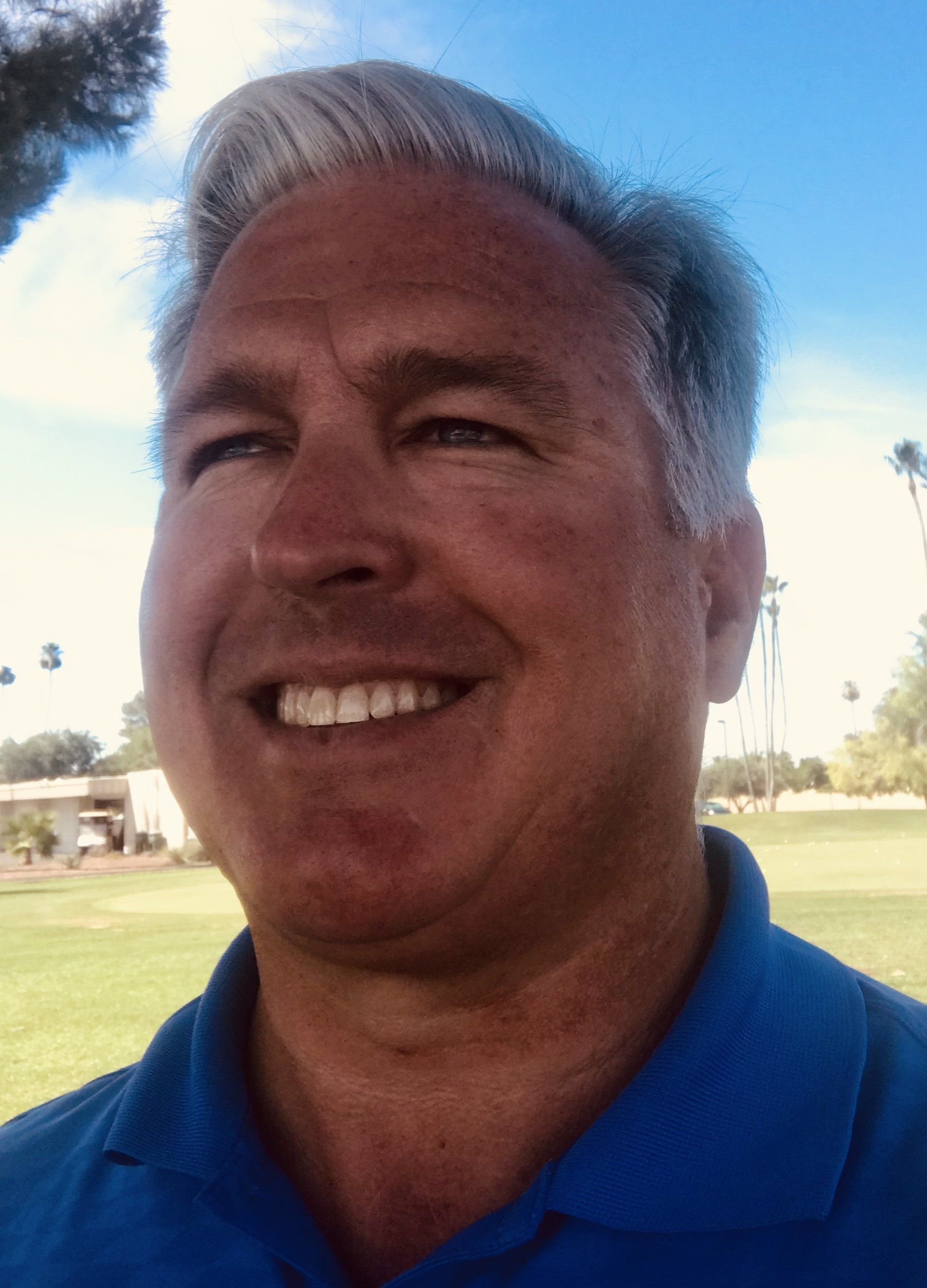 Man smiling outdoors, wearing a blue polo. Gray hair, golf course and sky in background.