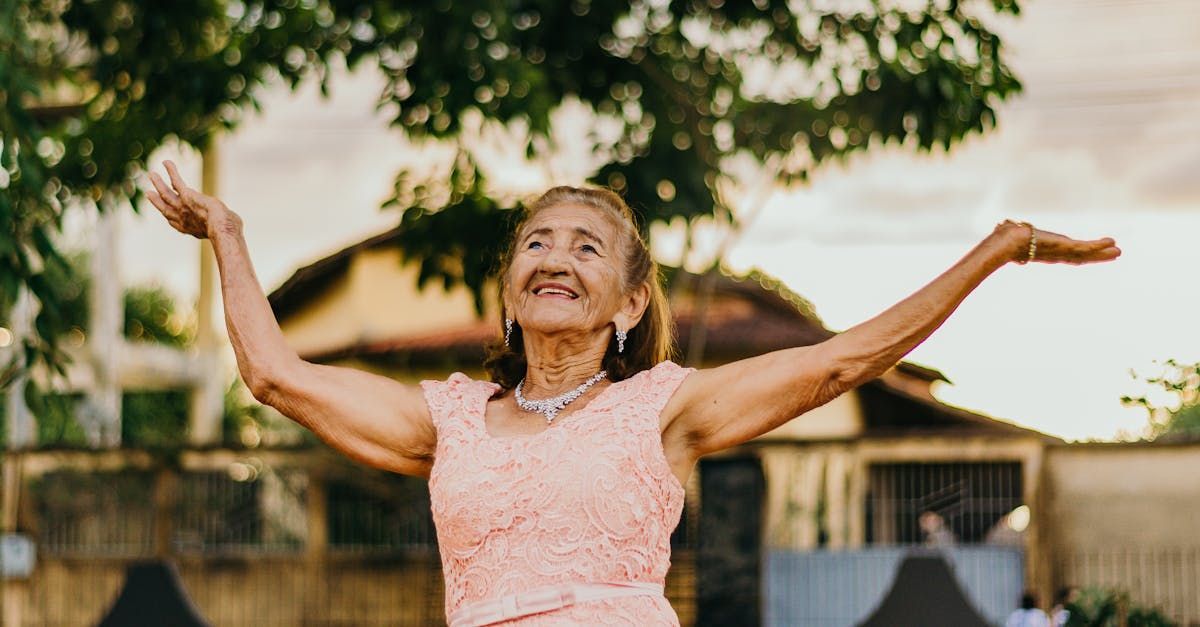 An elderly woman in a pink dress is dancing with her arms outstretched.