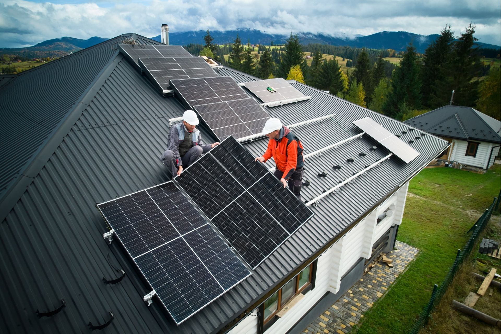 Two workers installing solar panels on a house roof with a mountain backdrop.