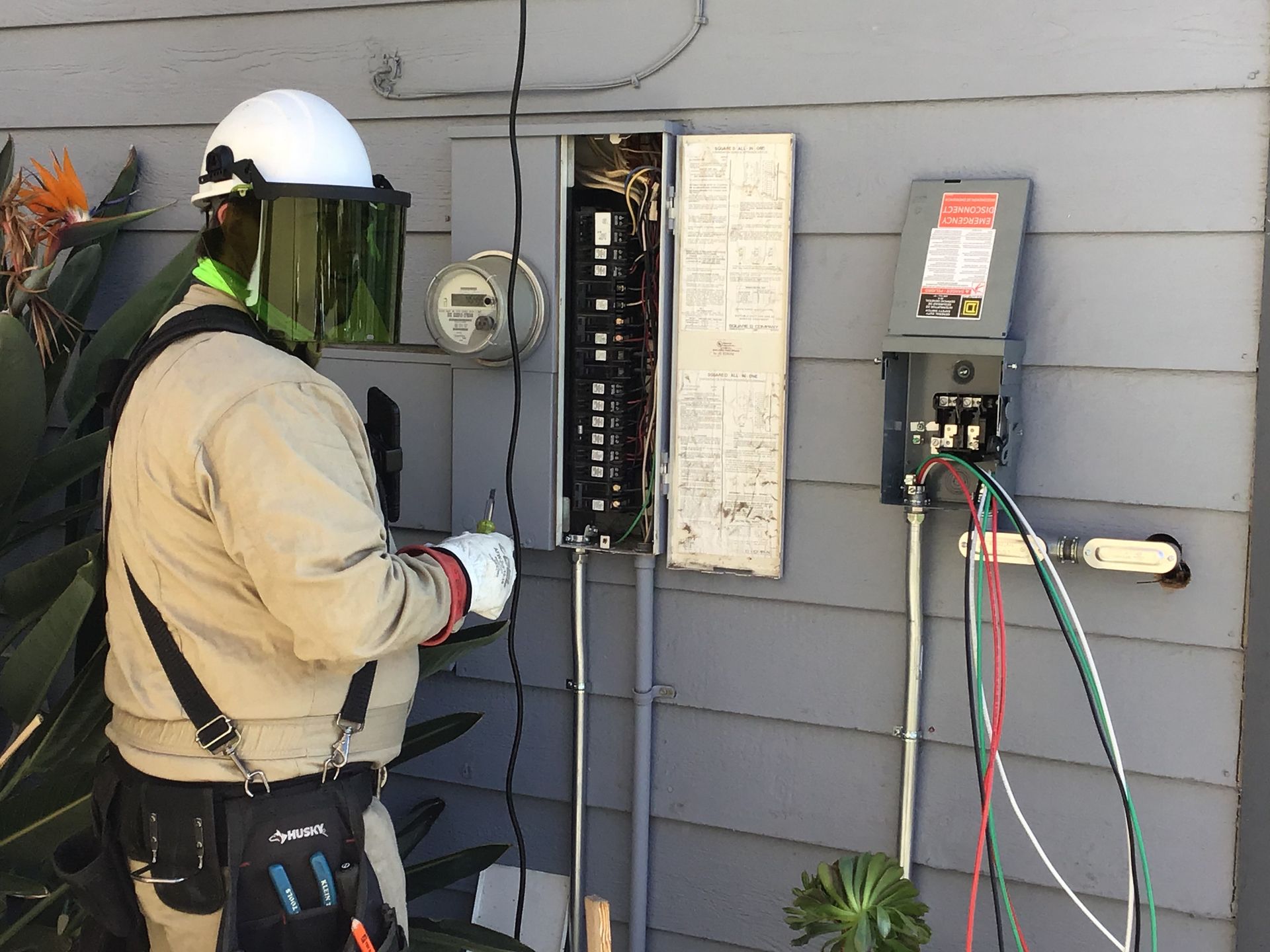 Electrician working on a circuit breaker panel outdoors; wearing protective gear.