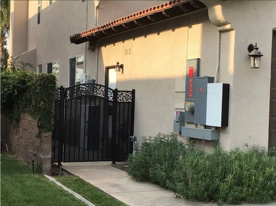 Black gate, walkway, and building with electrical boxes and a wall lantern in sunlight.
