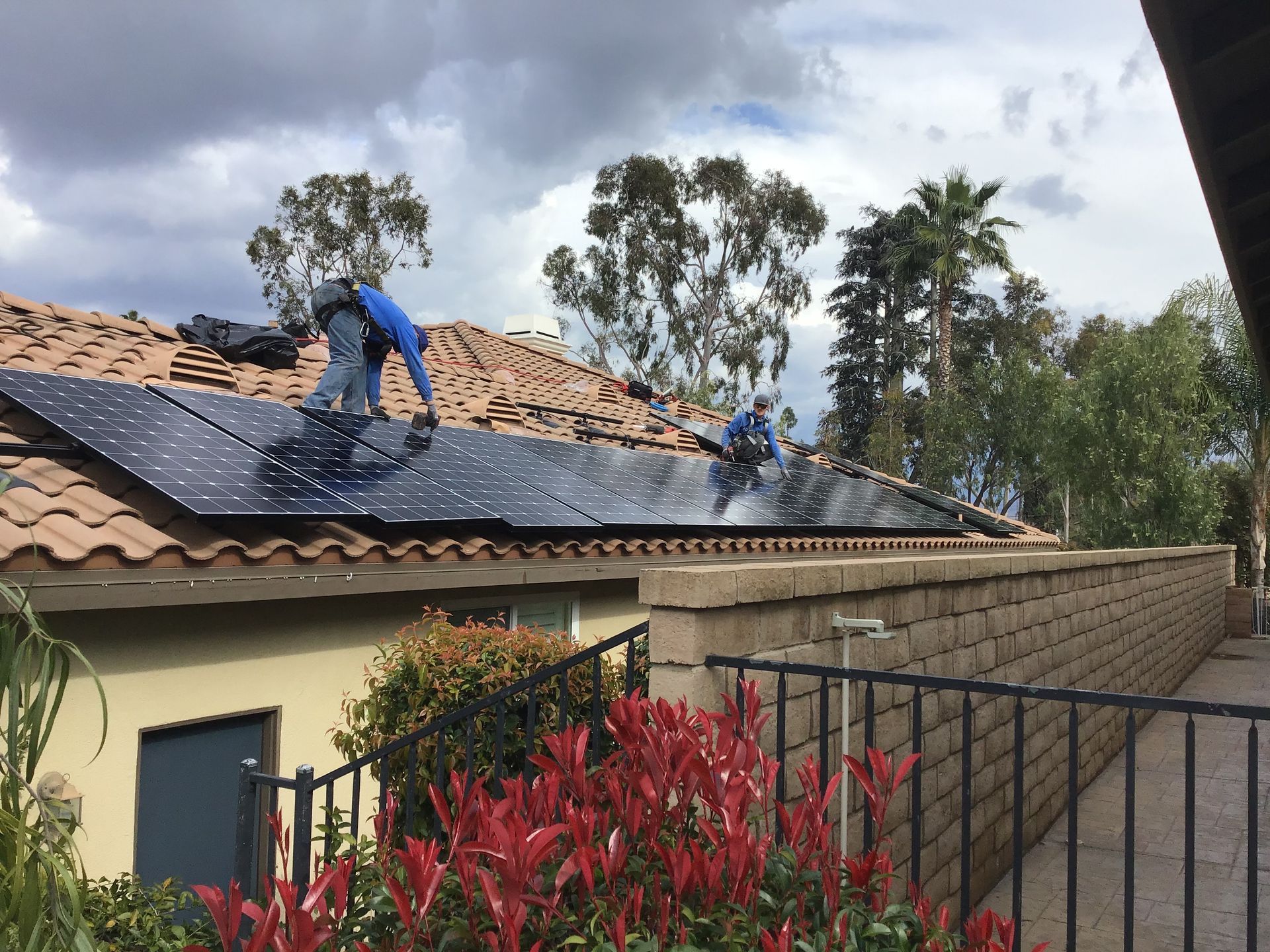 Workers installing solar panels on a tiled roof. Cloudy sky, red foliage, brick wall in the foreground.