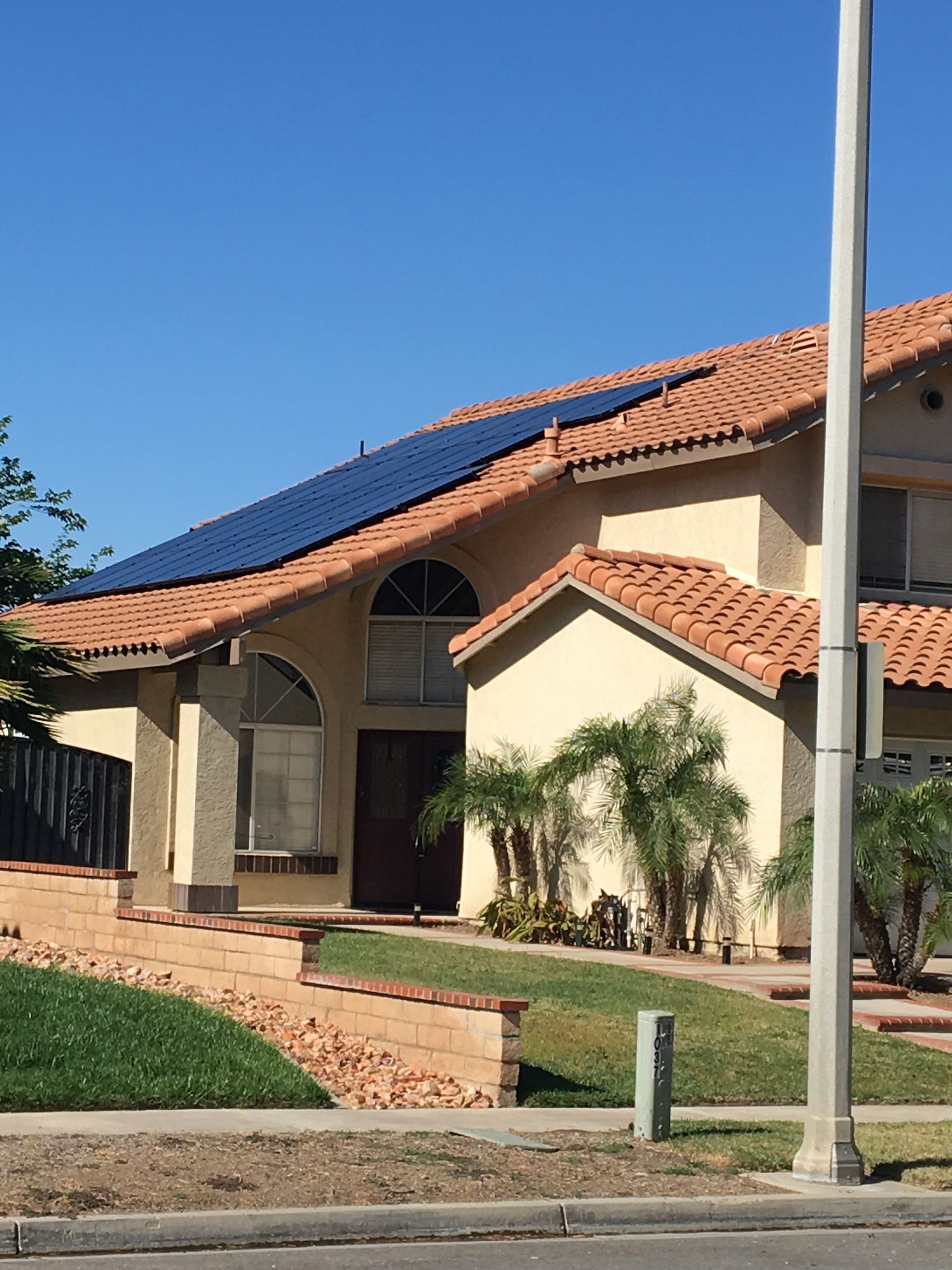 House with solar panels on the roof, stucco walls, and a terracotta tile roof, under a blue sky.