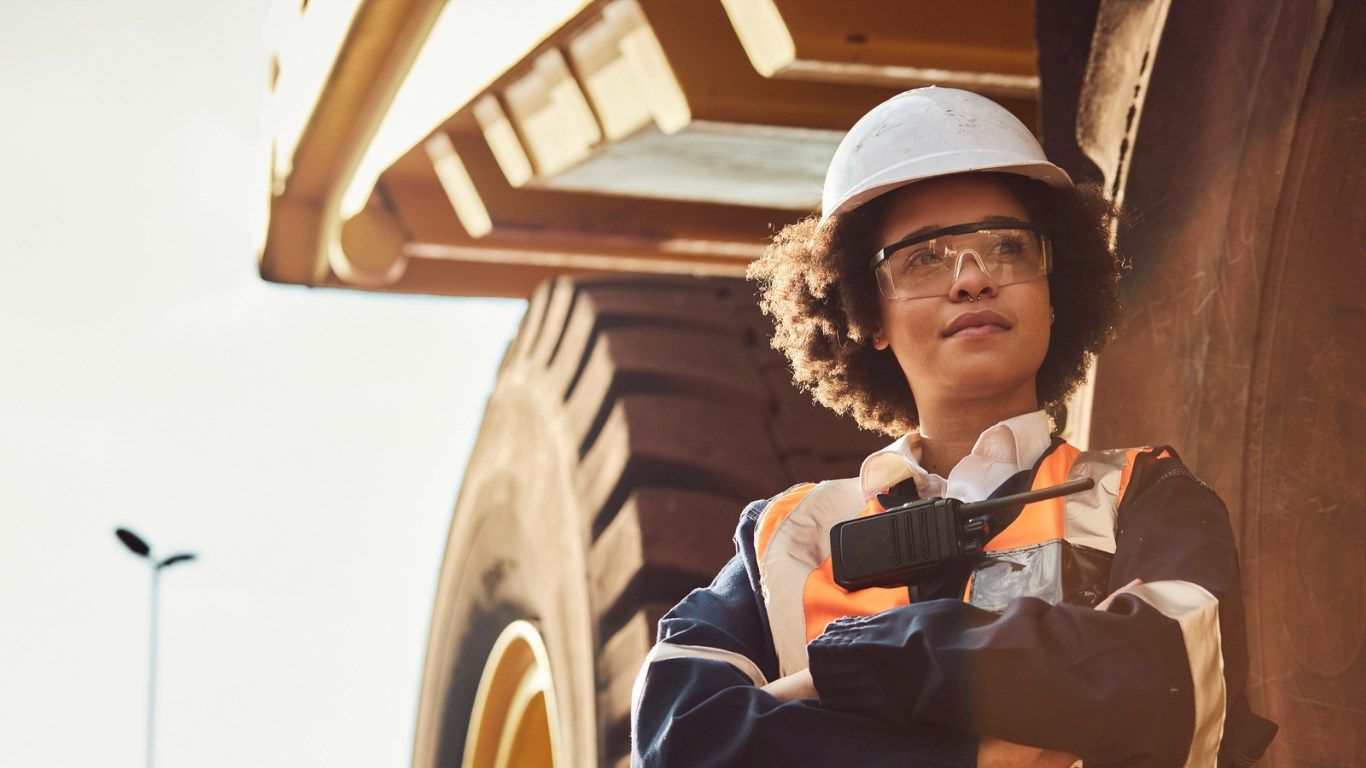 A woman wearing a hard hat , safety vest and glasses is standing in front of a building.