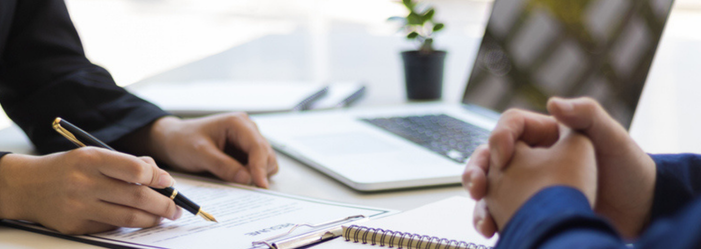 Two people are sitting at a table with their hands folded in front of a laptop computer.