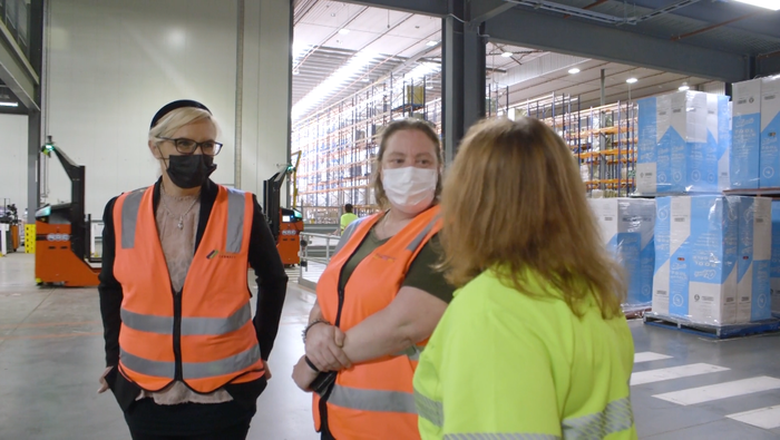 Three women wearing face masks are standing in a warehouse.